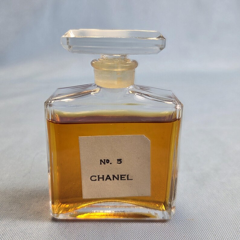 K&ouml;nnte beinhalten: Eine Vintage-Parf&uuml;mflasche Chanel No. 5. Die klare Glasflasche hat eine rechteckige Form mit einem durchsichtigen Verschluss. Auf dem Etikett steht "No. 5 CHANEL". Die Fl&uuml;ssigkeit im Inneren ist bernsteinfarben.