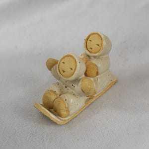 May include: A ceramic figurine depicts two figures in hooded winter suits riding a sled. The figures are beige with speckled details, and the sled is a light yellow. The figurine has a whimsical, handcrafted appearance.
