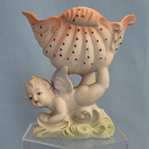 May include: A vintage porcelain figurine featuring a cherub supporting a seashell-shaped bowl. The cherub is in a reclining pose with small wings, and the shell is white with pink and brown accents. The base has green and gold details.