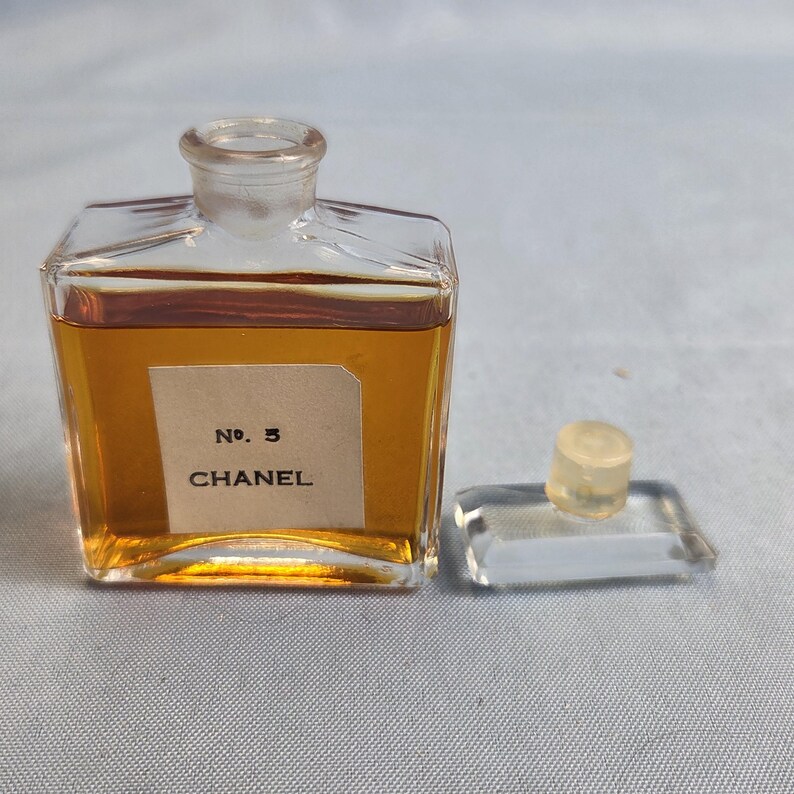 K&ouml;nnte beinhalten: Eine Vintage-Parf&uuml;mflasche Chanel No. 5. Die klare Glasflasche ist rechteckig mit abgerundeter Oberseite und enth&auml;lt bernsteinfarbene Fl&uuml;ssigkeit. Das Etikett lautet "No. 5 CHANEL". Der Verschluss ist separat und ruht auf einer klaren Glasbasis.