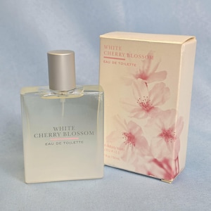 May include: A rectangular glass bottle of White Cherry Blossom eau de toilette with a silver cap, accompanied by its matching beige cardboard box featuring a pink cherry blossom floral print and the text 1.7 fl oz / 50 ml printed on the front.