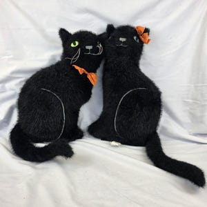 May include: Two plush black cats with fuzzy texture. The left cat has bright green eyes, white embroidered whiskers, and an orange ribbon around its neck. The right cat features an orange bow on its head and a grey nose. Both have long tails.