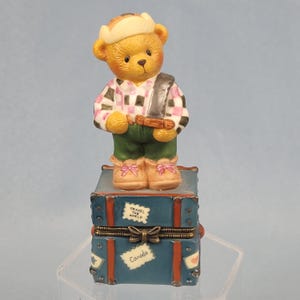 May include: A charming figurine of a teddy bear dressed in a plaid shirt, green pants, and boots, standing atop a blue travel trunk. The trunk has the words "Travel the World" and "Canada" on it. The bear has a hat and a strap across its chest.