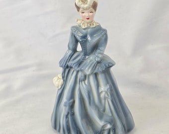May include: A porcelain figurine of a woman in a blue dress with white accents. The figurine has a white hat with gold trim and is holding a white purse. The dress has a ruffled collar and sleeves.