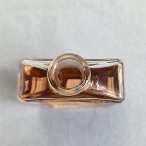 K&ouml;nnte beinhalten: Eine klare Parf&uuml;mflasche aus Glas mit rechteckiger Form und kreisf&ouml;rmiger &Ouml;ffnung. Die Flasche enth&auml;lt eine hell bernsteinfarbene Fl&uuml;ssigkeit, und das Glas reflektiert das Licht und erzeugt einen warmen Schein. Die Flasche ist leer.