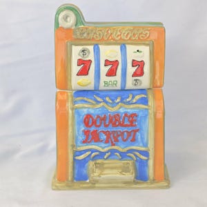 May include: A ceramic slot machine cookie jar with the word "Las Vegas" at the top. The slot machine has three reels with the number 7 and the word "BAR". The bottom of the jar reads "DOUBLE JACKPOT". The jar is painted in orange, blue, and gold.