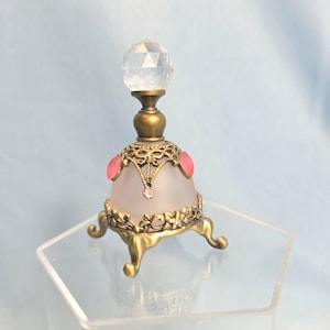 May include: A small perfume bottle with a frosted glass body, topped with a clear faceted crystal stopper. The base and neck feature an antique brass-toned metal filigree overlay, accented with small pink cabochons and a single pink rhinestone.