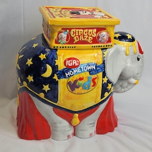 May include: A ceramic elephant-shaped cookie jar with a circus theme. The jar is painted in bright colors, including yellow, blue, and red. The lid is a box shape with the words "Circus Daze". The elephant has a blue cape with gold stars.