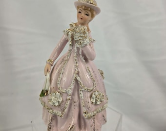 May include: A porcelain figurine of a woman in a pink dress and hat, holding a bouquet and a small purse. The dress is adorned with white floral accents and gold trim. The figurine has a vintage aesthetic.