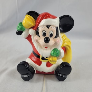 May include: A ceramic Mickey Mouse figurine dressed as Santa Claus. The figure is wearing a red Santa suit with white trim, a black belt, and a Santa hat. Mickey is holding a yellow bell and a green mitten. A yellow sack is behind him.