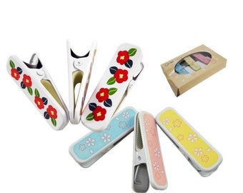 May include: Set of six colourful clothes pegs with floral designs. Three clothes pegs feature red camellia flowers and green leaves on a white background. The other three clothes pegs feature white flowers on a blue, pink, and yellow background.