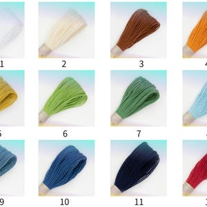 Puede incluir: Una colección de doce madejas de hilo de bordar numeradas en varios colores, incluyendo blanco, crema, marrón, naranja, amarillo, verde claro, verde oscuro, azul claro, azul turquesa, azul marino, azul oscuro y rojo, sujetas con una banda.