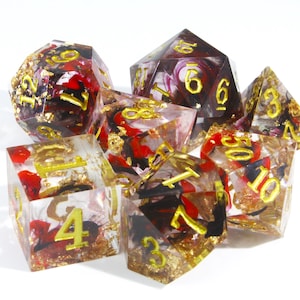 May include: A set of seven polyhedral dice for tabletop games. The dice are clear with red, black, and gold leaf inclusions. Each die has gold-coloured numbers.