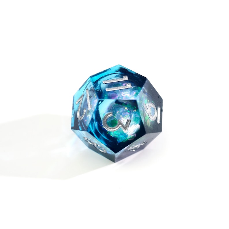 May include: A blue and teal D20 die with silver numbering. The die has a swirling, galaxy-like design inside the clear resin. The numbers are clearly visible, and the die is set against a white background.
