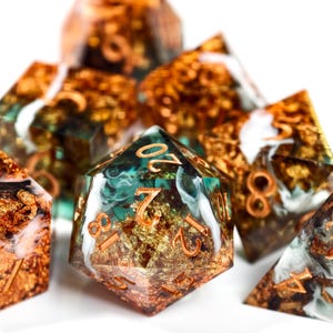 May include: A set of polyhedral dice with a distinctive design. The dice are transparent with gold foil and teal and white accents. The numbers are copper-coloured. The dice are for tabletop games.