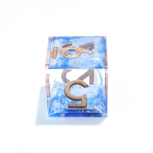 May include: A close-up of a clear, square die with the number 5 in gold. The die has a blue and white swirled interior. The die is set against a white background.