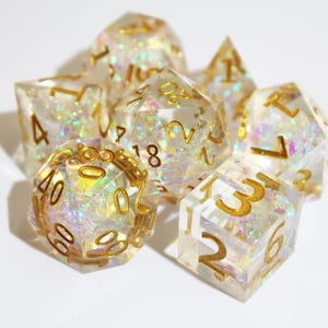 Op de afbeelding: Een set van zeven heldere, iriserende dobbelstenen met gouden cijfers. De dobbelstenen hebben verschillende polyhedrale vormen, waaronder een d4, d6, d8, d10, d12, d20 en een percentieldobbelsteen. Deze dobbelstenen zijn ontworpen voor tabletop-rollenspellen.