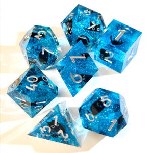 Può includere: Un set di sette dadi poliedrici. Ogni dado è blu trasparente con glitter e sagome di gatti neri. I numeri sono argentati. I dadi includono un D4, D6, D8, D10, D12, D20 e un dado percentuale.