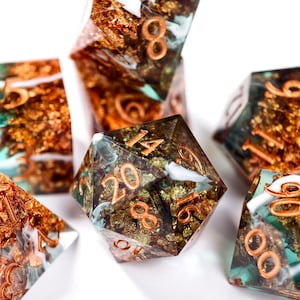 Op de afbeelding: Close-up van een set polyhedrale dobbelstenen. De dobbelstenen zijn transparant met goudfolie en koperkleurige cijfers. De dobbelstenen hebben turquoise en witte accenten. De dobbelstenen zijn voor tabletopspellen.