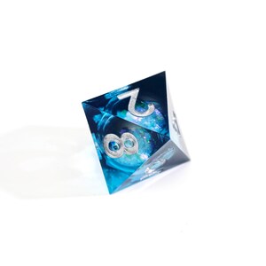 May include: A blue and clear resin D20 die with silver numbering. The die has a galaxy-like interior with the number 8 and 2 visible. The die is a polyhedral shape, commonly used in tabletop role-playing games.