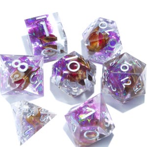 Puede incluir: Juego de siete dados poliédricos para juegos de rol. Cada dado es transparente con purpurina morada, flores secas y numeración plateada. El juego incluye un d4, d6, d8, d10, d12, d20 y un dado de porcentaje.