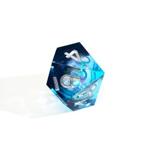 May include: A blue and clear D20 die with silver numbering. The die has a swirling, galaxy-like interior with glitter. The numbers are in a metallic silver color. The die is set against a white background.