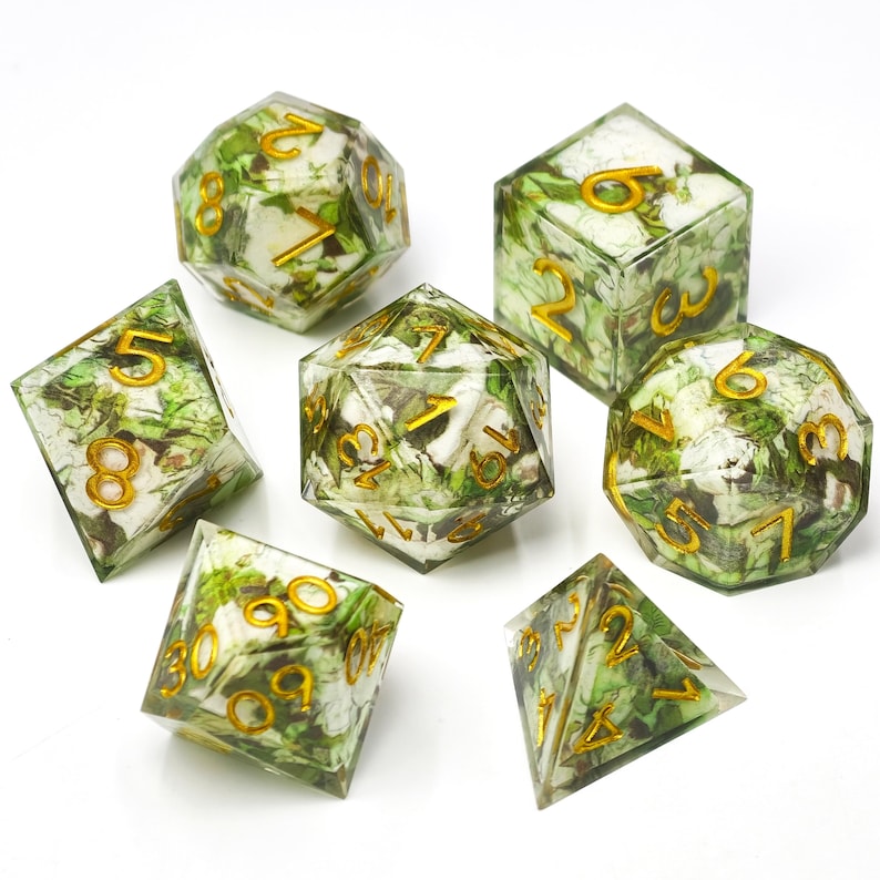 K&ouml;nnte beinhalten: Ein Set aus sieben polyedrischen W&uuml;rfeln aus klarem Harz mit eingebetteten wei&szlig;en und gr&uuml;nen floralen Elementen. Jeder W&uuml;rfel hat scharfe Kanten und gravierte Zahlen, die mit einem metallisch goldenen Pigment gef&uuml;llt sind, darunter ein W20, W12, W10, W8, W6, W4 und ein Prozentw&uuml;rfel.