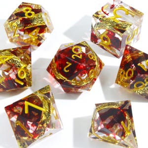 May include: Set of seven polyhedral dice with a clear resin exterior, red interior, and gold-colored numbers and decorative accents. The dice are designed for tabletop role-playing games.