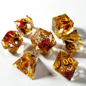 May include: A set of seven polyhedral dice. Each die is transparent with gold-coloured numbers and embedded with red flower petals and gold flakes. The dice are arranged on a white surface.