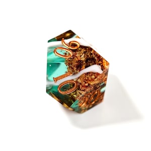 Op de afbeelding: Een polyhedrale dobbelsteen met een turquoise en gouden binnenkant, met koperkleurige cijfers. De dobbelsteen heeft een wit, wervelend ontwerp en staat tegen een witte achtergrond. De cijfers zijn duidelijk zichtbaar.