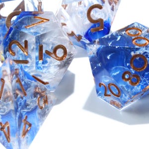 May include: Close-up of several polyhedral dice. The dice are clear with blue and white swirls, and bronze-colored numbers. The dice are used for tabletop role-playing games.