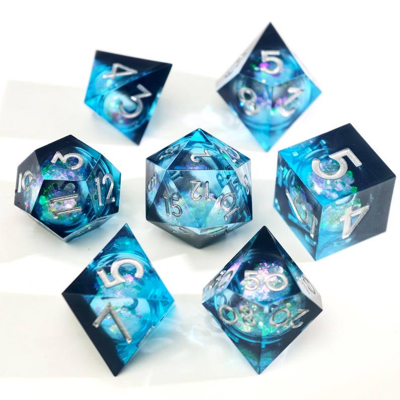 May include: Set of seven polyhedral dice with a blue and black color scheme. The dice have a swirling, galaxy-like design inside, with silver numbering. The dice are arranged on a white surface.