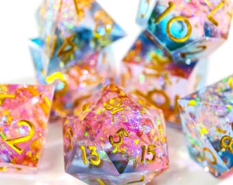 May include: A collection of polyhedral dice in various shapes, featuring a colorful, glittery design. The dice are primarily pink, blue, and gold, with gold numbering. These dice are suitable for tabletop games.