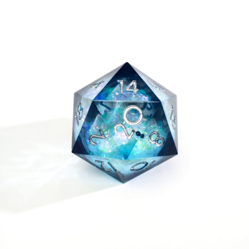 May include: A clear, multi-faceted D20 die with a blue and turquoise interior, featuring silver numbering. The die has a celestial design with glitter and small, dark blue accents, perfect for tabletop gaming.