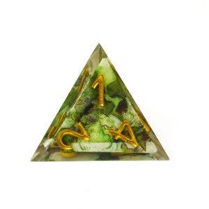 K&ouml;nnte beinhalten: Ein vierseitiger Tetraeder-W&uuml;rfel aus klarem Harz mit einem eingebetteten gr&uuml;n-braunen Blumenstoffmuster. Der W&uuml;rfel hat erhabene goldfarbene Ziffern auf seinen Fl&auml;chen, wobei die Zahlen eins, zwei und vier deutlich zu erkennen sind.