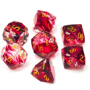Peut inclure: Un ensemble de sept dés polyédriques en résine rouge translucide avec des inclusions sombres tourbillonnantes et des chiffres gravés de couleur dorée. Le lot comprend diverses formes géométriques pour les jeux de société, avec des bords nets.