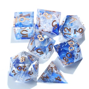 May include: A set of seven polyhedral dice with a blue and white marbled design. The dice have gold-colored numbers and are made of a clear resin material. The set includes a D4, D6, D8, D10, D12, D20, and a percentile die.