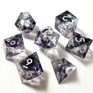 May include: Set of seven polyhedral dice for tabletop games. The dice are clear with a dark, swirling interior design and silver numbering. Includes a d4, d6, d8, d10, d12, d20, and a percentile die.