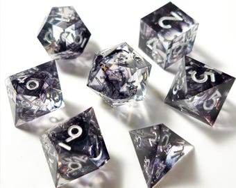May include: Set of seven polyhedral dice for tabletop games. The dice are clear with a dark, swirling interior design and silver numbering. Includes a d4, d6, d8, d10, d12, d20, and a percentile die.
