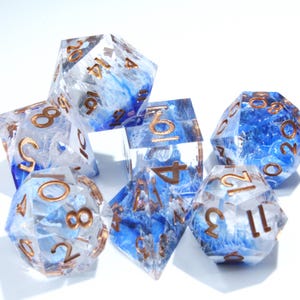 May include: A set of seven polyhedral dice for tabletop games. The dice are clear with blue and white swirls, and gold-colored numbers. The dice include a D4, D6, D8, D10, D12, D20, and a percentile die.