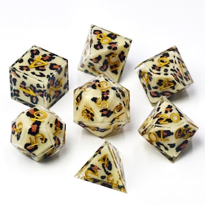 May include: A set of seven polyhedral dice featuring an off-white base with a black and brown leopard print pattern and recessed gold-coloured numbers. The collection includes a d4, d6, d8, d10, d12, d20, and a percentile die with clear resin edges.