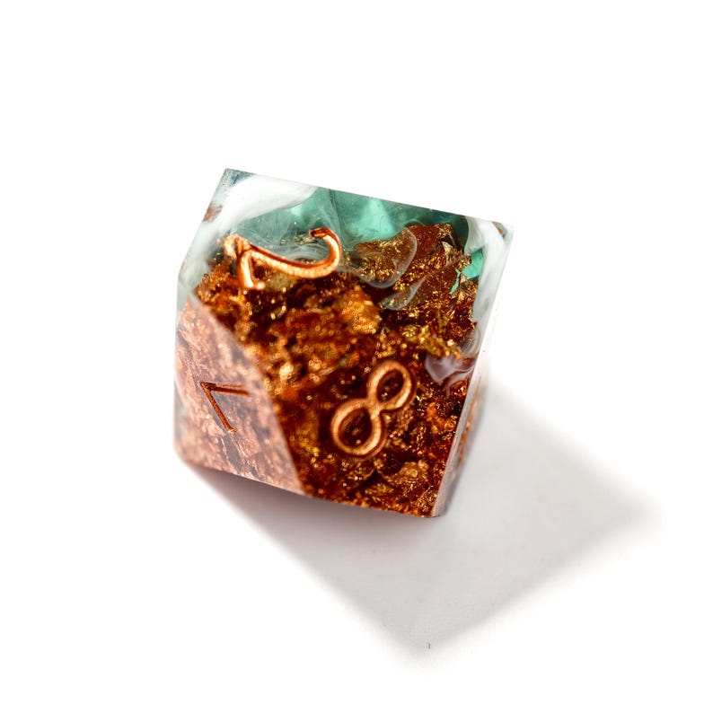 Op de afbeelding: Een handgemaakte D20 dobbelsteen van heldere hars, met koperkleurige insluitsels en turquoise accenten. De cijfers 7 en 8 zijn koperkleurig. Ontworpen voor tabletop games.