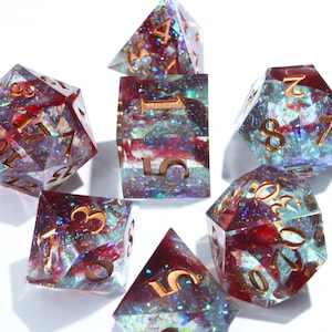 Puede incluir: Un conjunto de siete dados poliédricos. Los dados son transparentes con purpurina iridiscente y detalles rojos. Los números son dorados. Los dados están dispuestos sobre una superficie blanca.