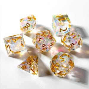 May include: A set of seven clear, iridescent polyhedral dice with gold-colored numbers. The dice are arranged on a white surface, with each die showing a different face. The dice are designed for tabletop role-playing games.