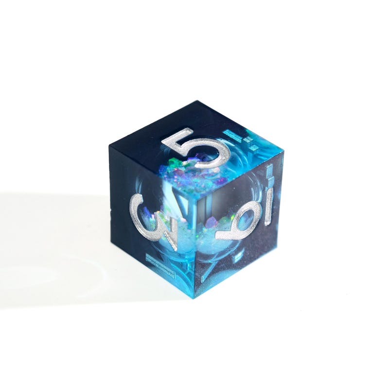 May include: A clear resin die with a dark blue and turquoise interior, featuring silver numerals. The number 5 is visible on top, with other numbers on the sides. The die has a shimmering, galaxy-like effect.