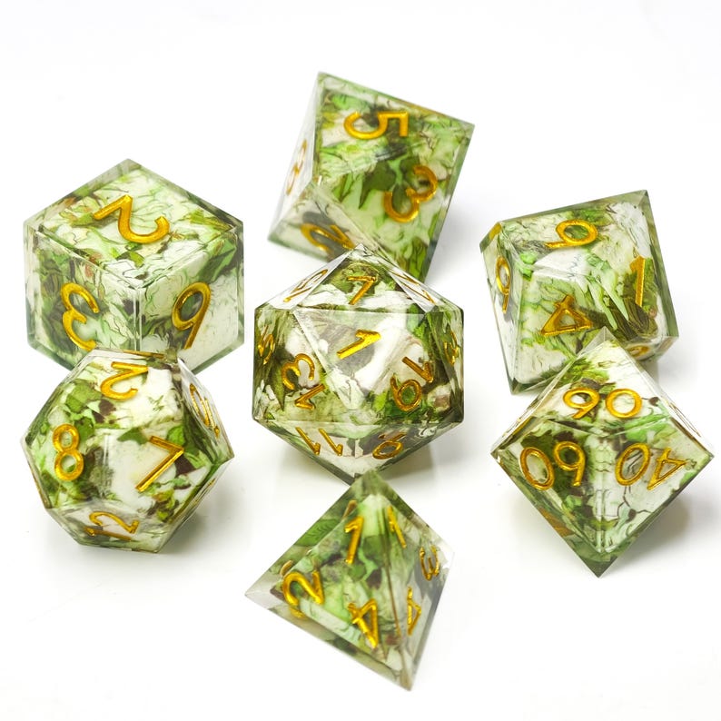 K&ouml;nnte beinhalten: Ein Set aus sieben polyedrischen W&uuml;rfeln aus klarem Harz mit eingebetteten gr&uuml;nen Bl&auml;ttern und wei&szlig;en floralen Elementen. Jeder W&uuml;rfel hat scharfe Kanten und goldfarbene, eingravierte Zahlen auf den Seiten, darunter ein W20, W12, W10, W8, W6, W4 und ein Prozentw&uuml;rfel.
