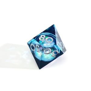 May include: A D20 dice with a blue and teal interior, and silver numbers. The dice is made of resin and has a clear, transparent appearance. The numbers are clearly visible and the dice is set against a white background.