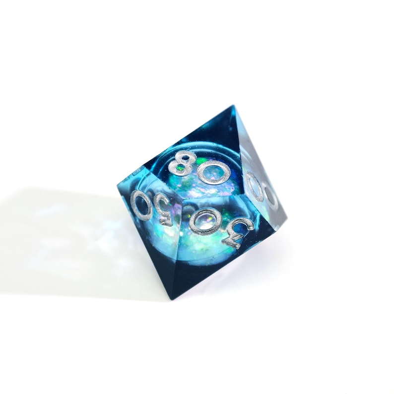 May include: A D20 dice with a blue and teal interior, and silver numbers. The dice is made of resin and has a clear, transparent appearance. The numbers are clearly visible and the dice is set against a white background.