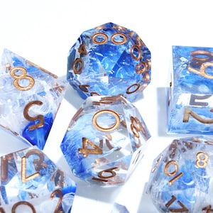 May include: Set of polyhedral dice with a blue and white marbled design. The dice have gold-colored numbers and edges. The dice are made of a clear resin material.