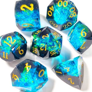 May include: Seven polyhedral dice with a dark, translucent base and vibrant blue and green internal designs. Each die features gold-colored numbers. The dice are arranged on a white surface, showcasing their multifaceted shapes.